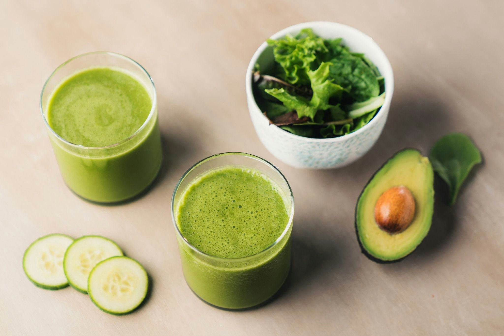 two glasses of green smoothie adjacent to sliced, fresh avocado, kale, and sliced cucumber