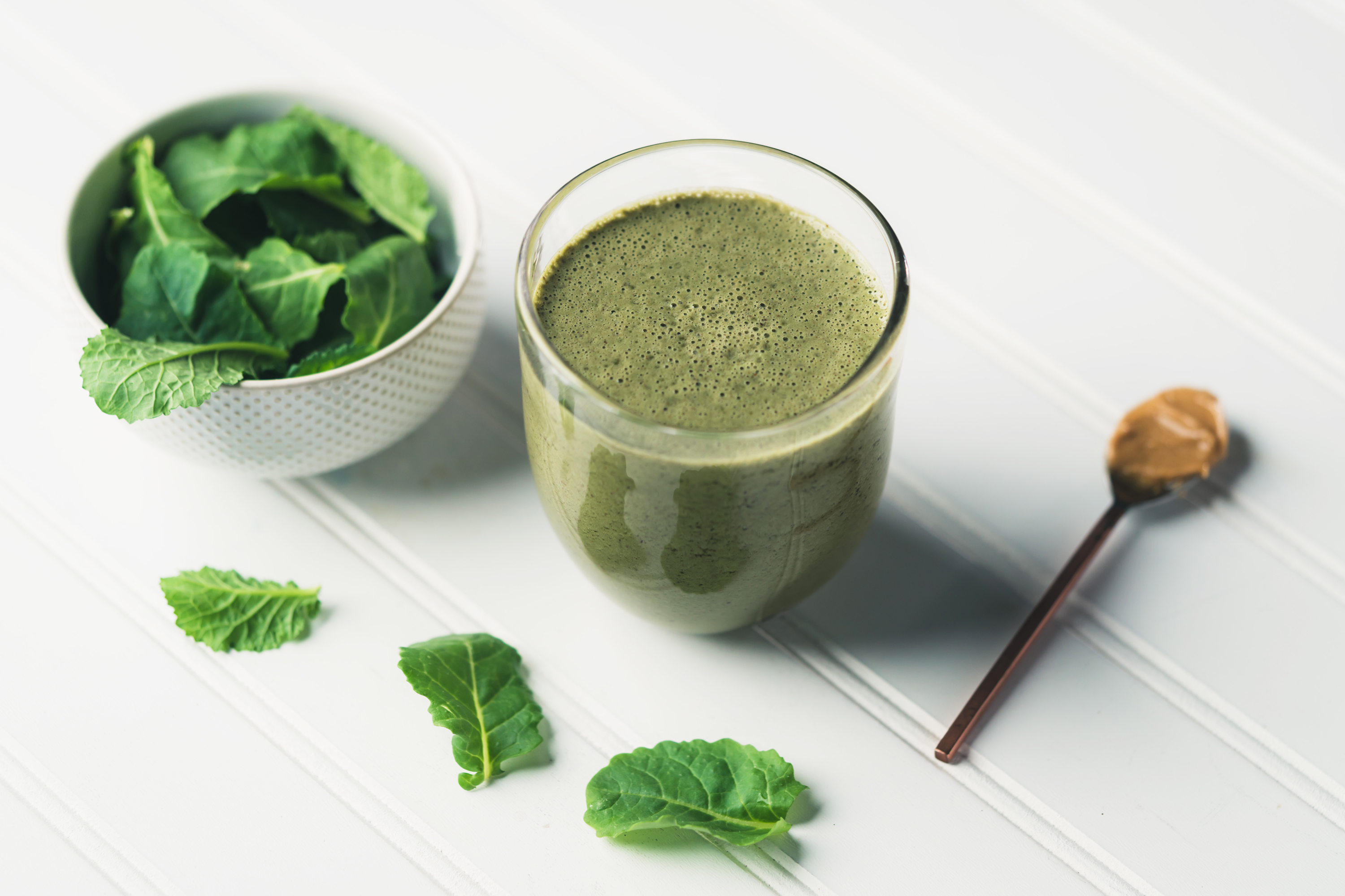 a short glass filled with a green smoothie next to a cup of fresh spinach and a spoonful of cashew butter