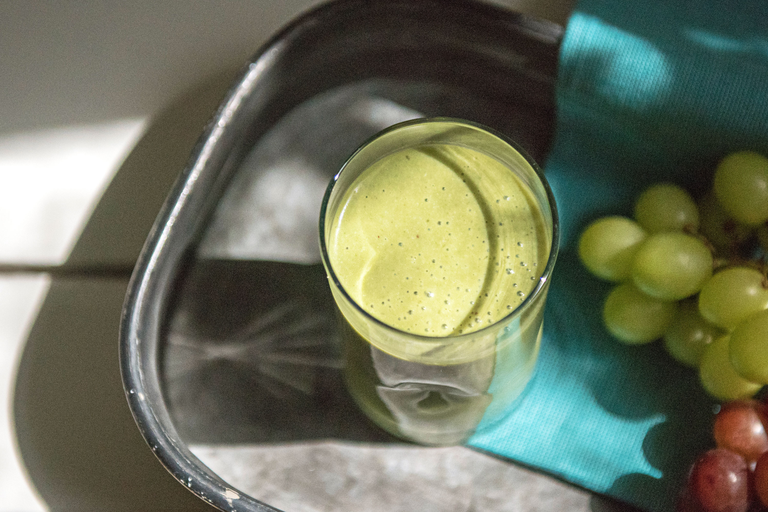 green smoothie in a tall glass on a tray next to fresh green and purple grapes