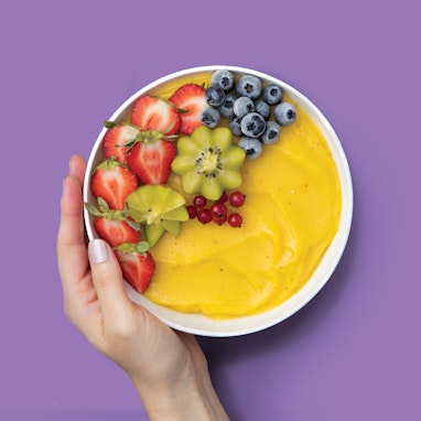 hand holding bowl of yellow stone fruit smoothie with fresh berries and kiwi