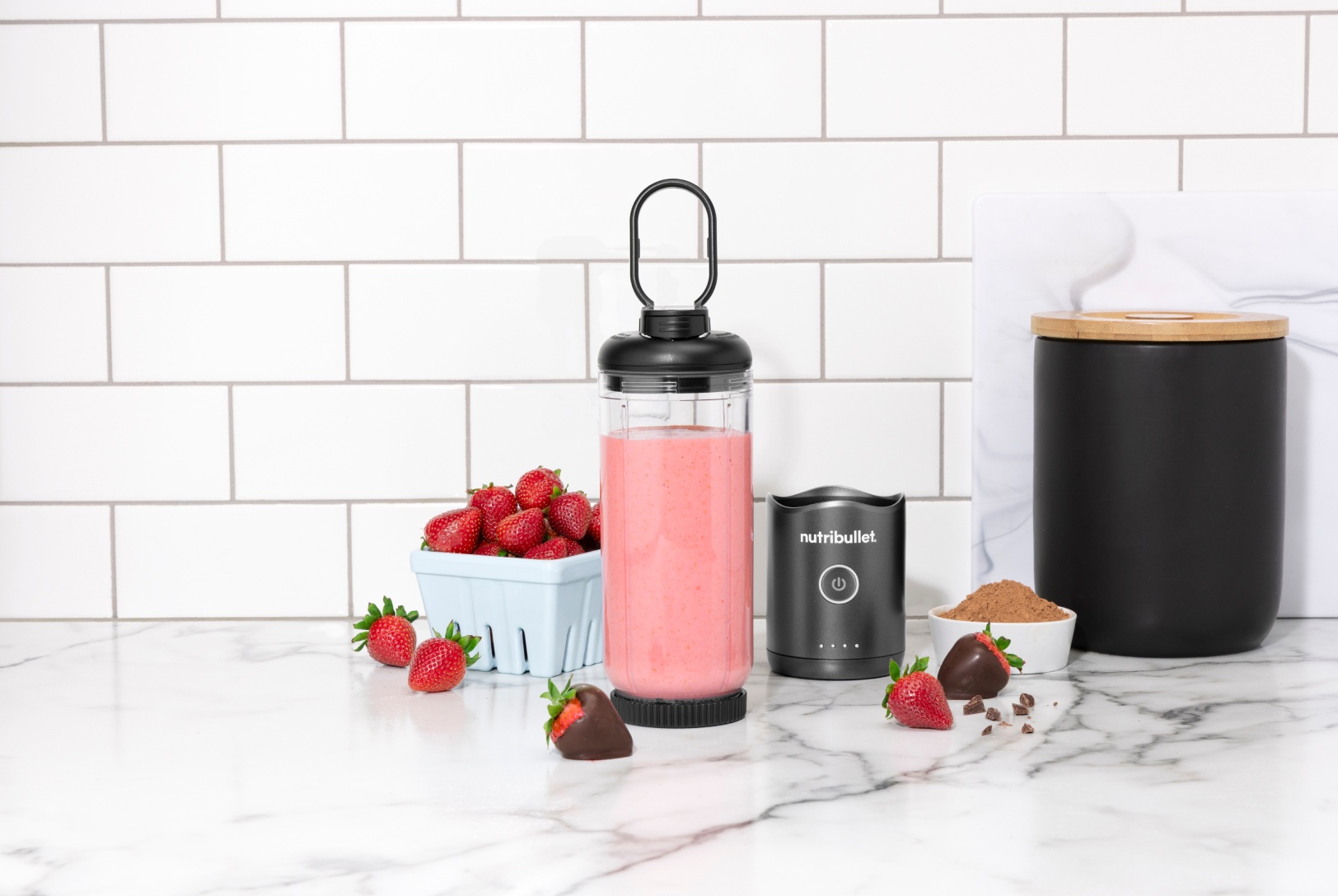 pink smoothie in a portable blender placed next to a pint of fresh strawberries and chocolate covered strawberries