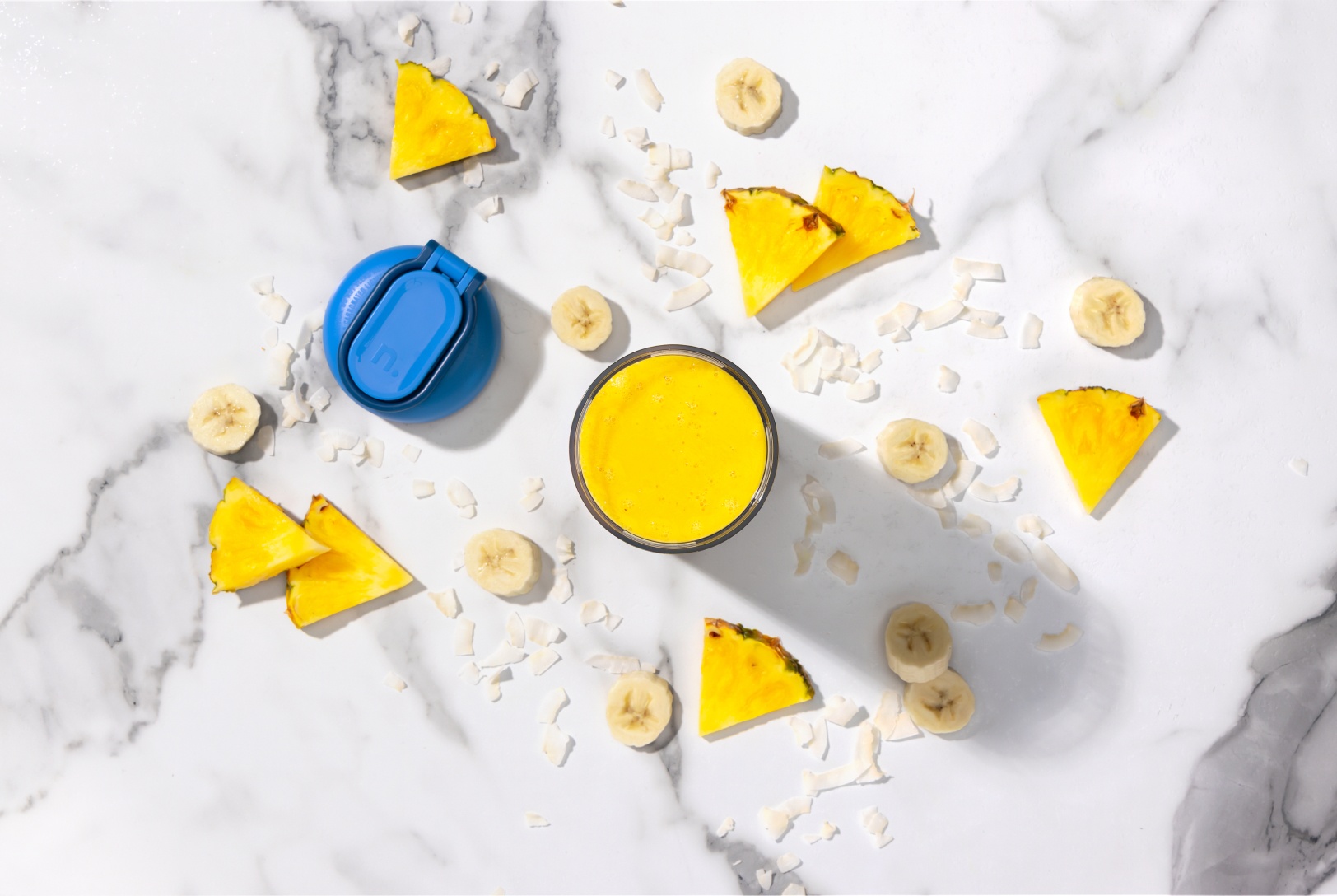 bird's eye view of turmeric smoothie surrounded by fresh pineapple, banana, and a blue portable blender lid