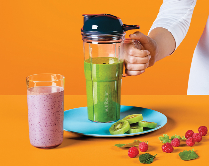 Cup of purple smoothie and travel cup of green smoothie with fruits on blue plate on orange background.