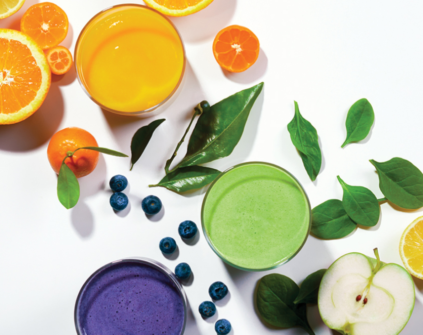Three glasses of orange, green, and purple juices with fruits and greens on white background.