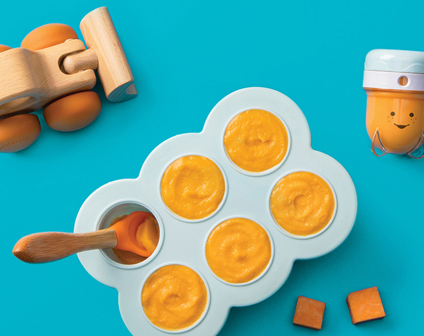 Silicone tray filled with orange baby food, baby bullet accessory and a toy on a blue background.