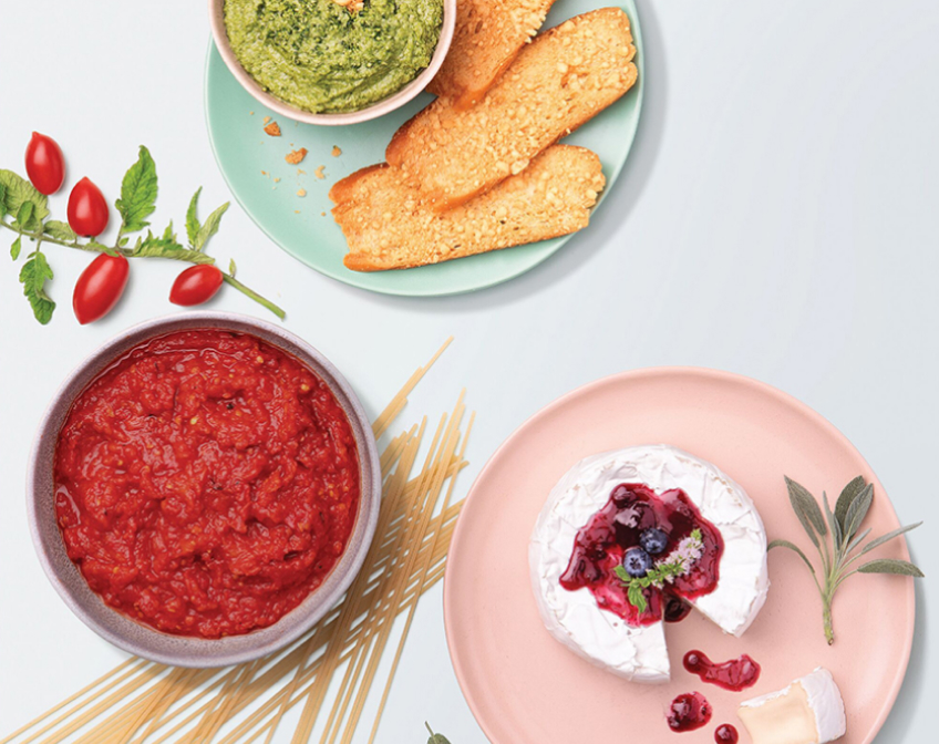 Blue plate of green dip and crackers, purple bowl of red sauce, pink plate of white cheese next to uncooked pasta, cherry tomatoes on white background
