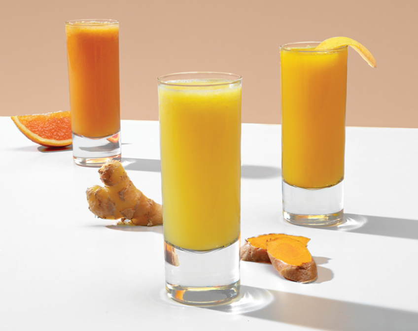 Three glasses of yellow and orange juices with ginger root and fruits on white surface.