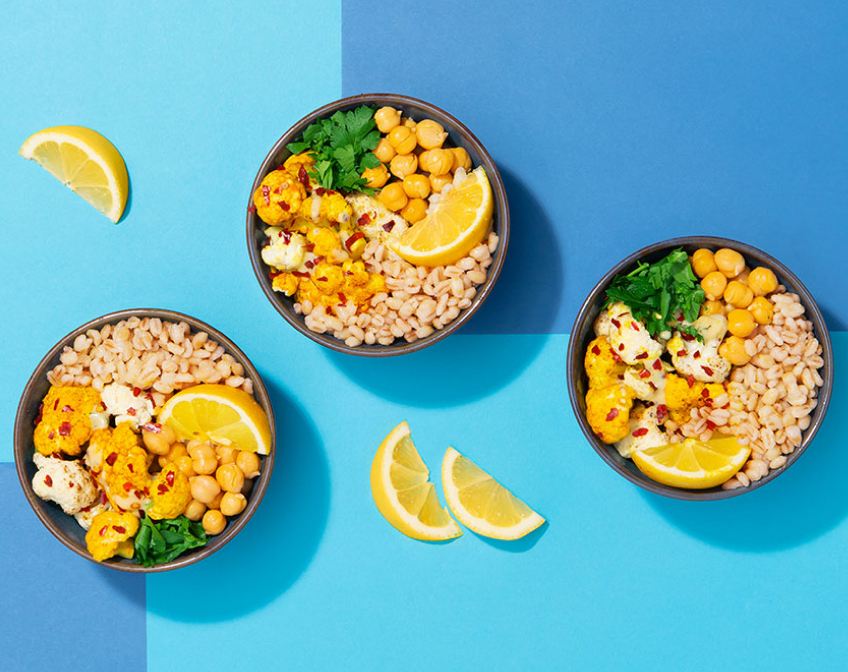 Three bowls of cauliflower, chickpea and barley salad on a blue surface with lemons.