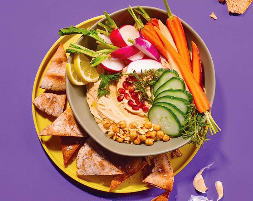 A tan bowl filled with veggies and hummus on top of a yellow plate of chips against a purple background