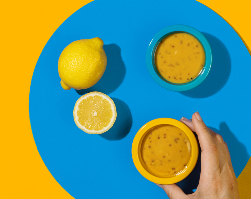 Blue and yellow bowls of dressing with hand grabbing one with lemons on blue and yellow background.