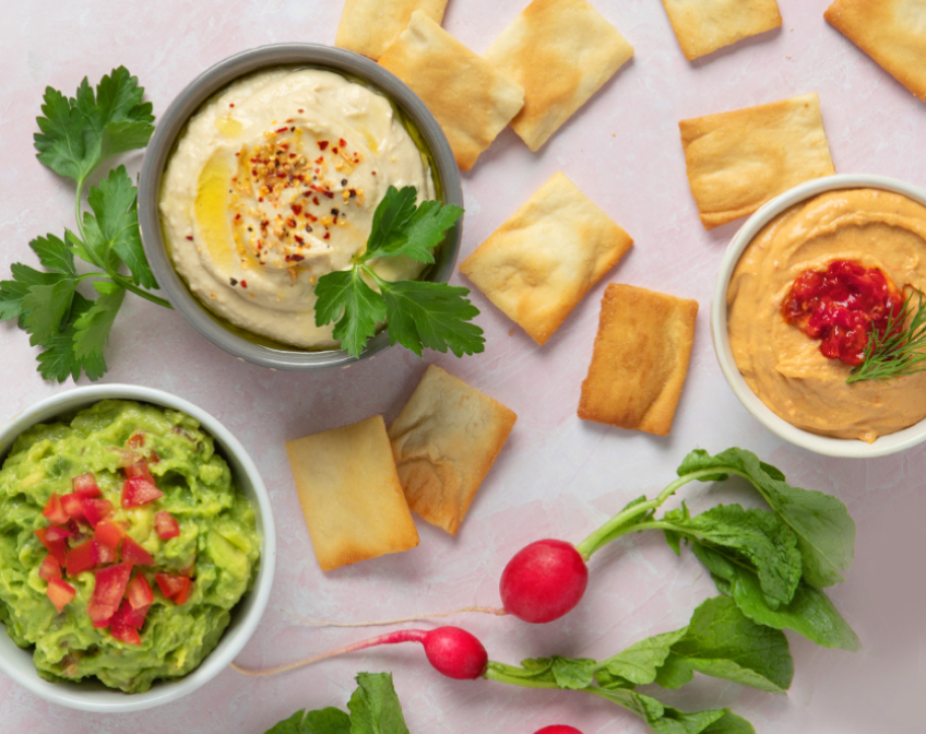 Three bowls of dips on a white surface with crackers and greenery.