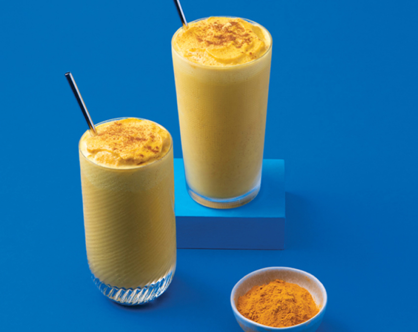 Two glasses of yellow smoothie with metal straws with a bowl of seasoning on blue background.