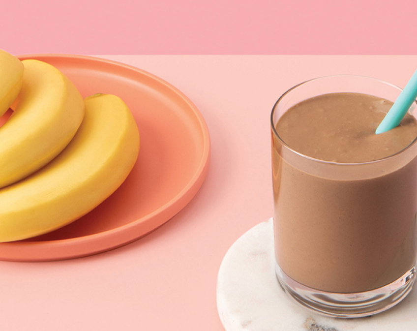 Glass of brown smoothie with blue straw and an orange plate of bananas on pink background.