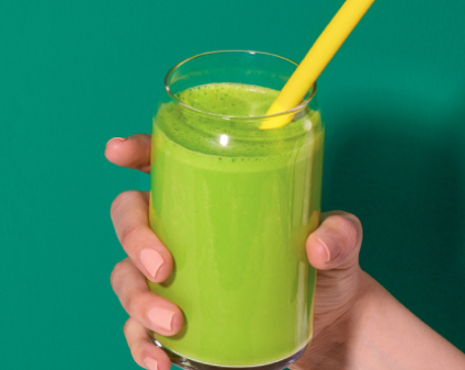 Hand holding a glass of green juice with yellow straw on green background.