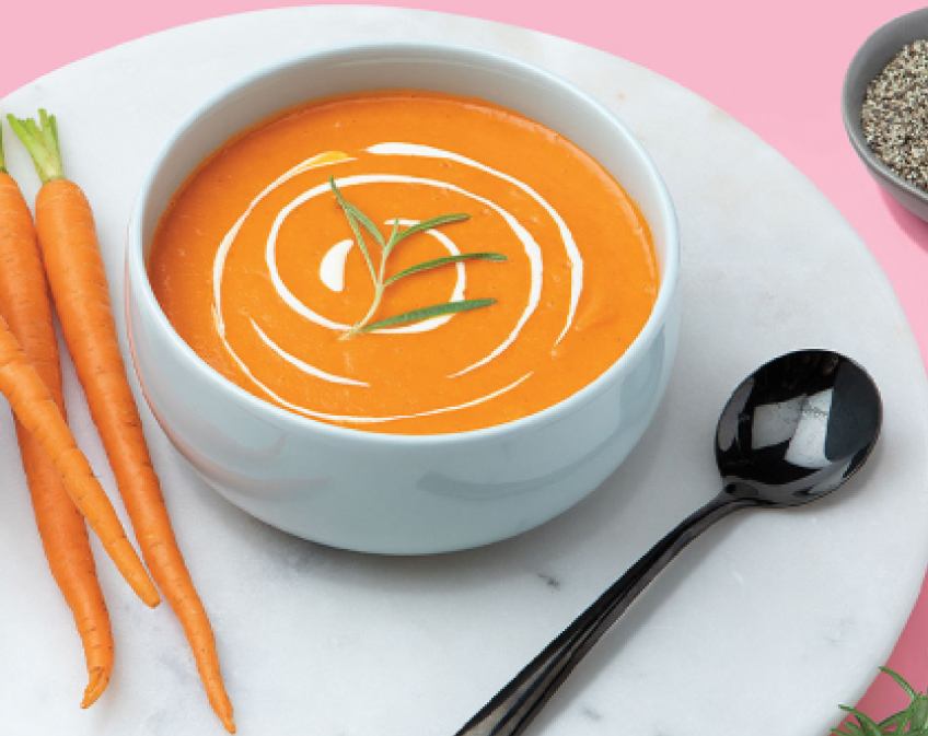 White bowl of orange soup with green sprig, carrots, and black spoon on white plate on pink surface.