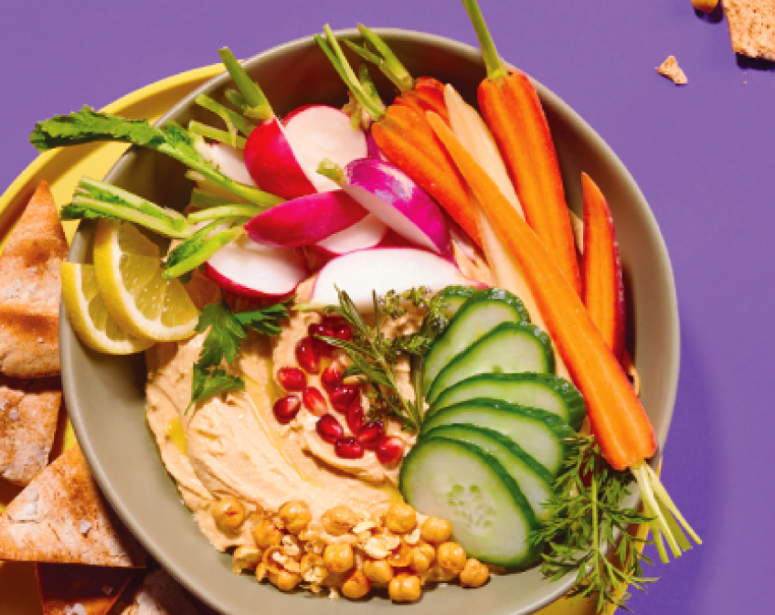 Bowl of hummus, beans, and colorful vegetables on yellow plate with pita on purple surface.