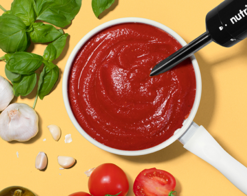 Red sauce in white saucepan with green leaves, tomatoes, and garlic on yellow surface.